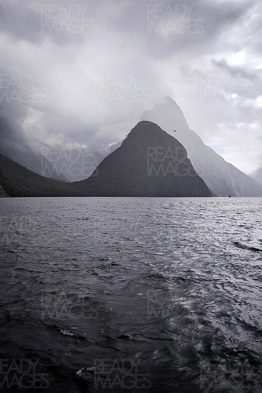 Black and white fine art image taken on a moody dramatic weather, looking at the ocean and mountains of Milford Sound in the background