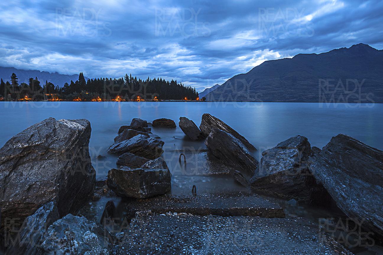 Long exposure seascape image with large rocks, blurry water, mountains and cloudy blue sky, taken during blue hour