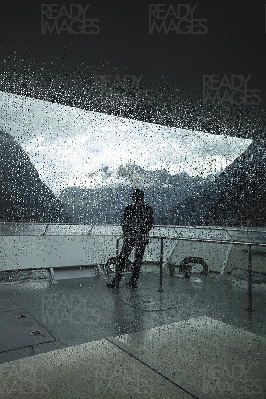 Image of a tourist looking at Fiordland National Park Mountains from the boat, taken on a dramatic and rainy day