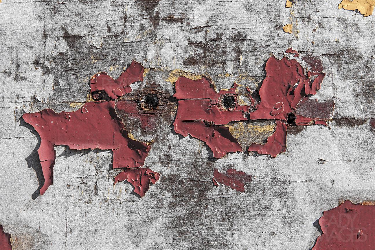 Close-up image of a weathered, chipped red paint peeling off old damaged timber with holes